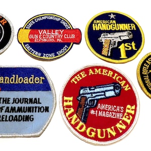 May include: A collection of embroidered patches featuring firearm-related logos and text. Patches include "Ruger Firearms," "American Handgunner," and "Handloader." The patches are in various shapes and colors, with detailed stitching.