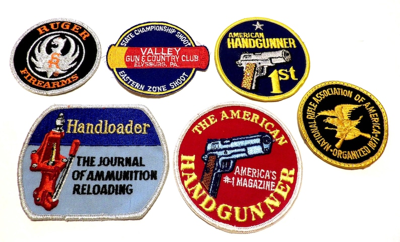 May include: A collection of embroidered patches featuring firearm-related logos and text. Patches include "Ruger Firearms," "American Handgunner," and "Handloader." The patches are in various shapes and colors, with detailed stitching.