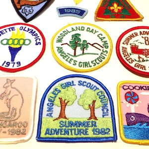 May include: A collection of vintage Girl Scout patches with various designs and text. Patches include "Cadette Olympics 1979", "Woodland Day Camp", "Summer Adventures '81", "Kangaroo Day - 1982", "Angeles Girl Scout Council Summer Adventure 1982", and "Cookies".