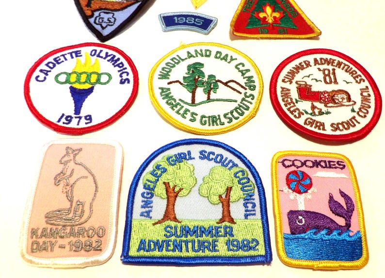 May include: A collection of vintage Girl Scout patches with various designs and text. Patches include "Cadette Olympics 1979", "Woodland Day Camp", "Summer Adventures '81", "Kangaroo Day - 1982", "Angeles Girl Scout Council Summer Adventure 1982", and "Cookies".