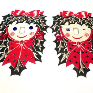 Puede incluir: Dos cubiertas de interruptor de luz de Navidad vintage con la cara de una niña con una corona de acebo y un lazo rojo. Las cubiertas tienen un fondo blanco con contornos negros y detalles de purpurina roja y azul.