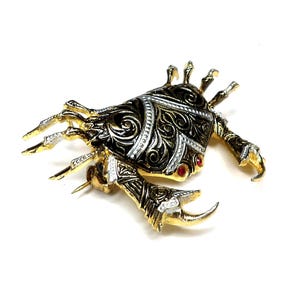 Puede incluir: Broche de cangrejo dorado con intrincados detalles de filigrana negra y plateada. El cuerpo del cangrejo presenta detalles rojos y está sobre un fondo blanco. Las pinzas y las patas son doradas con reflejos plateados.