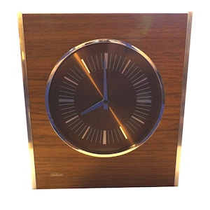 Könnte beinhalten: Eine braune Holzrahmenuhr mit silbernem Zifferblatt und schwarzen Zeigern. Das Zifferblatt der Uhr hat einen silbernen Rand und der Markenname "Sandoz" ist in der rechten unteren Ecke aufgedruckt.