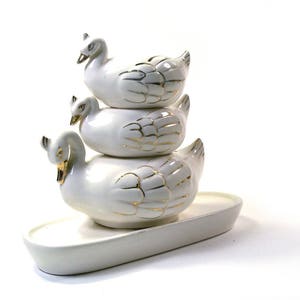 Può includere: Un set di tre figurine di cigno in ceramica bianca impilate l'una sull'altra. I cigni hanno dettagli dorati e sono seduti su un vassoio in ceramica bianca.
