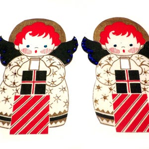 Puede incluir: Dos adornos navideños vintage con forma de ángeles que sostienen regalos. Los ángeles son blancos con estrellas doradas y alas azules. Tienen el pelo rojo y llevan vestidos blancos con ribetes negros. Los regalos son de rayas rojas y blancas con cajas negras y blancas.