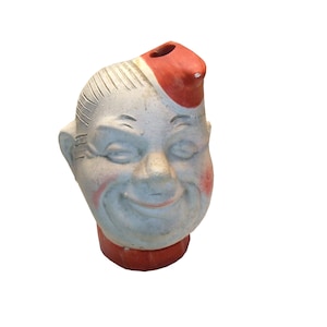May include: A ceramic figurine of a smiling man's head with a red hat. The figurine is painted in shades of gray, red, and white.