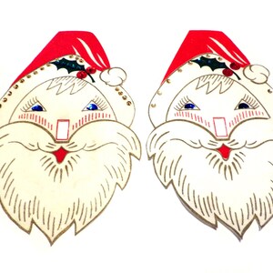 Puede incluir: Dos recortes de caras de Papá Noel vintage. Cada Papá Noel tiene un gorro rojo, ojos azules y barba blanca. Los sombreros están decorados con acebo y detalles dorados. Una decoración navideña festiva.