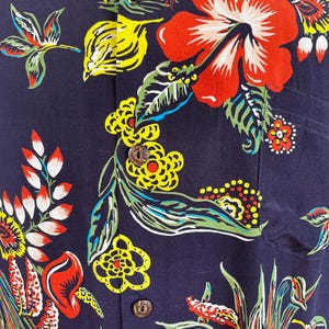 Peut inclure: Chemise bleu marine &agrave; motifs floraux. Le motif pr&eacute;sente de grandes fleurs d'hibiscus rouges, des feuilles jaunes et vertes et de plus petites fleurs color&eacute;es. La chemise a des boutons en bois et une poche poitrine.