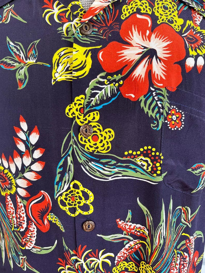 Peut inclure: Chemise bleu marine &agrave; motifs floraux. Le motif pr&eacute;sente de grandes fleurs d'hibiscus rouges, des feuilles jaunes et vertes et de plus petites fleurs color&eacute;es. La chemise a des boutons en bois et une poche poitrine.