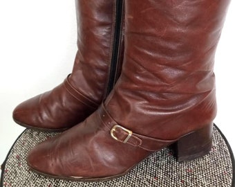 May include: A pair of knee-high brown leather boots. The boots have a pointed toe, a small heel, and a buckle detail around the ankle. A black zipper runs along the side of each boot.