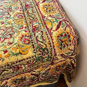 May include: A yellow, red, and green patterned textile with a floral design. The fabric is draped over a surface, showcasing the intricate details of the print. The textile features a border with a repeating paisley pattern.