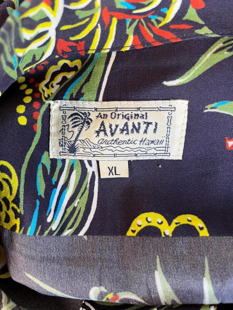 Peut inclure: Gros plan sur une chemise hawa&iuml;enne bleu fonc&eacute; avec un motif floral color&eacute;. La chemise porte une &eacute;tiquette indiquant "An Original Avanti authentic Hawaii" et la taille "XL". Le motif de la chemise comprend des touches de rouge, jaune, vert et bleu.