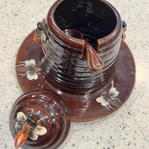 May include: A brown ceramic honey pot with a matching lid and saucer. The pot has a dark blue interior and is decorated with bee-shaped accents and painted bee illustrations. The lid features a bee-shaped handle.