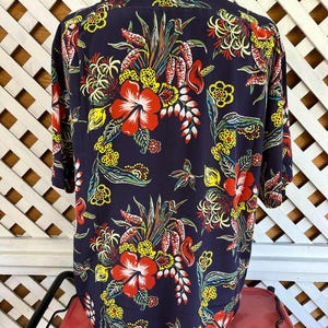 Peut inclure: Une chemise bleu marine &agrave; boutons avec un motif floral vibrant. Le design pr&eacute;sente de grandes fleurs d'hibiscus rouges, des accents jaunes et divers feuillages tropicaux. La chemise a des manches courtes et un col classique.