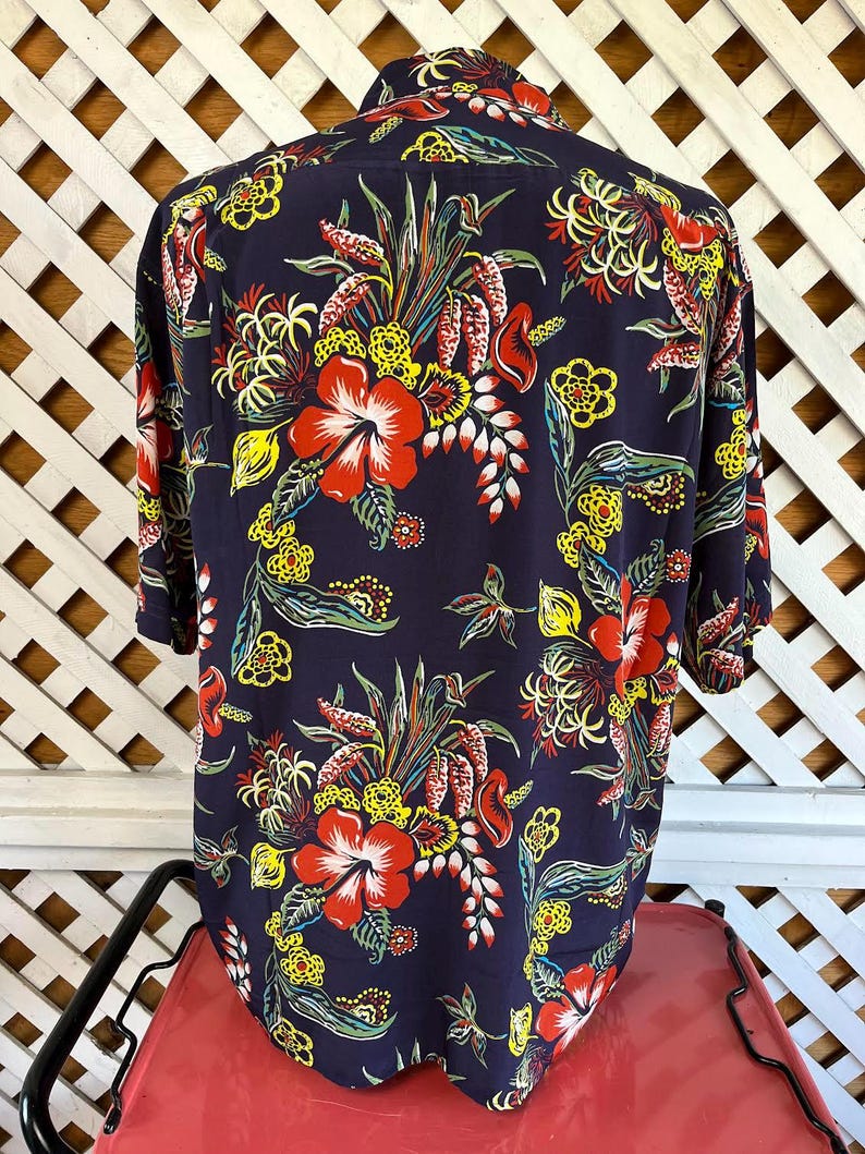 Peut inclure: Une chemise bleu marine &agrave; boutons avec un motif floral vibrant. Le design pr&eacute;sente de grandes fleurs d'hibiscus rouges, des accents jaunes et divers feuillages tropicaux. La chemise a des manches courtes et un col classique.