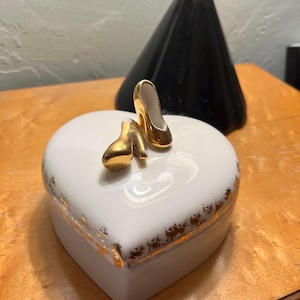 May include: A white heart-shaped trinket box with gold trim and two miniature gold high heels on top. The box is on a wooden surface, with a black lamp in the background.