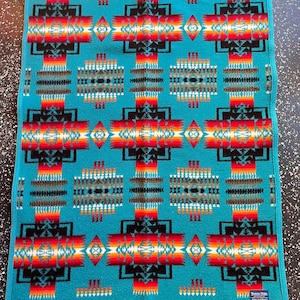 May include: A turquoise blanket with a Southwestern-inspired geometric pattern. The design features repeating crosses and diamond shapes in red, orange, black, and white. The blanket has a rectangular shape and a finished edge.