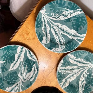 May include: Three round ceramic plates with a teal and white tropical leaf pattern. The plates are arranged on a wooden surface, showcasing the detailed botanical design. The plates are a decorative element for the dining table.