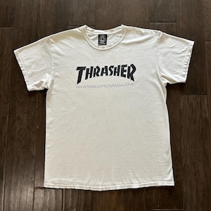 以下が含まれることがあります： 黒い太字の「THRASHER」の文字と、その下に小さな黒い文字で「SKATEBOARD MAGAZINE」と書かれた白いTシャツ。Tシャツは濃い木製の表面に平らに置かれています。