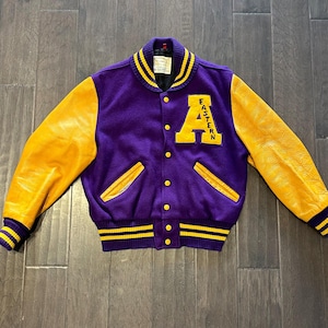 May include: A vintage purple and gold letterman jacket with the word "EASTERN" on the front. The jacket has gold leather sleeves, a purple body, and gold and black striped cuffs and collar. The jacket has a button-up front and two front pockets.