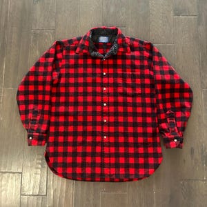 Pode incluir: Uma camisa de manga comprida com botões e um padrão de xadrez vermelho e preto. A camisa tem um forro de colarinho preto, um bolso no peito no lado esquerdo e uma bainha curva. Uma etiqueta da marca Pendleton é visível no colarinho.