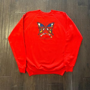 Puede incluir: Una sudadera roja con un diseño de mariposa en azul, amarillo y blanco. El gráfico de la mariposa está centrado en la parte delantera, encima del texto "RASATHON". La sudadera está hecha de un material suave.