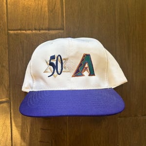 May include: White baseball cap with a blue brim. The cap features the number "50" and the letter "A" embroidered on the front. The "A" is in teal, purple, and brown. The cap is likely a sports team souvenir.