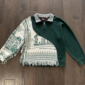 May include: Green long-sleeve sweatshirt with a gray collar and a quarter-zip closure. The left side features a tapestry design with fringe, depicting a courthouse and the text "Hillsboro City Library".