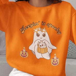 May include: Orange sweatshirt with a graphic of a friendly ghost wearing a flower crown and holding a pumpkin basket. The text "Keepin' it spooky" is arched above the ghost. Two jack-o'-lanterns with candles are on either side of the ghost.