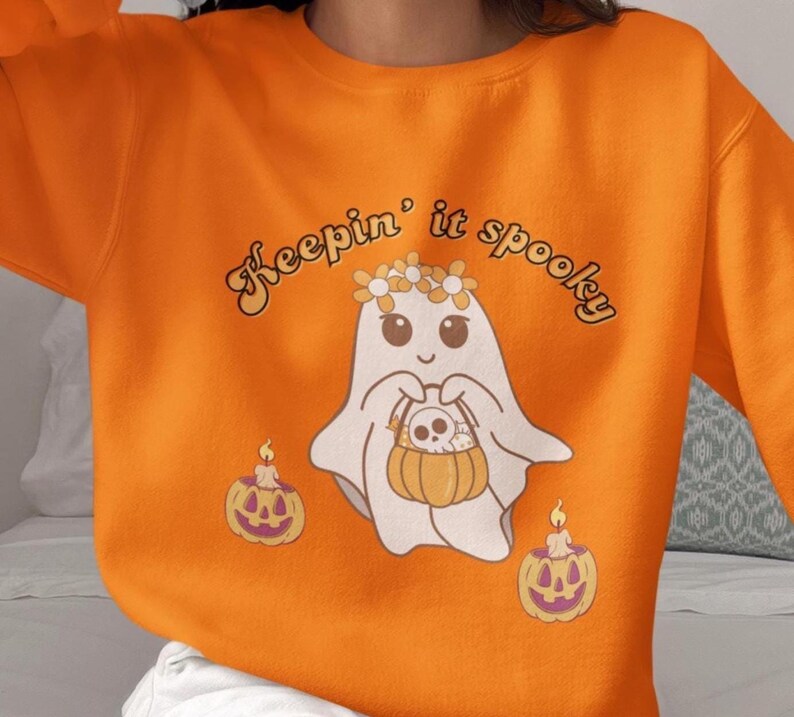 May include: Orange sweatshirt with a graphic of a friendly ghost wearing a flower crown and holding a pumpkin basket. The text "Keepin' it spooky" is arched above the ghost. Two jack-o'-lanterns with candles are on either side of the ghost.