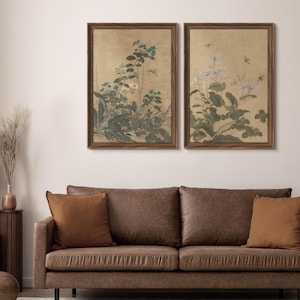 May include: Two framed botanical prints with a vintage aesthetic. The artwork features detailed illustrations of flowers and foliage in shades of green, blue, and white against a beige background. The frames are a dark brown color, and the prints are displayed above a brown sofa.