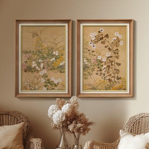 May include: Two framed floral art prints. Each print features a botanical illustration of white flowers, green leaves, and tall grasses against a tan background. The frames are light brown with a white inner border.
