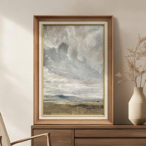 May include: A framed landscape painting with a cloudy sky and a field. The artwork features a wooden frame with a gold inner border. The painting's color palette includes shades of gray, brown, and green, creating a serene and natural aesthetic.