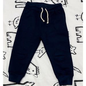 Puede incluir: Pantalones deportivos azul marino con cintura con cordón y parches en las rodillas. Los pantalones están hechos de un material suave y son adecuados para ropa casual. Los pantalones se muestran sobre un fondo blanco estampado.