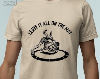 Può includere: T-shirt beige con grafica nera di due lottatori su un tappeto. Il testo "LEAVE IT ALL ON THE MAT" è arcuato sopra i lottatori. La maglietta ha girocollo e maniche corte.