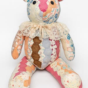May include: A handmade patchwork teddy bear with a cream lace collar and a cream ribbon. The bear's head, arms, and legs are made of colorful fabric patches in shades of pink, blue, orange, and brown. The bear has black button eyes.