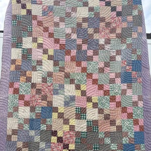 May include: A patchwork quilt featuring a grid of small multi-colored fabric squares in shades of pink, blue, green, yellow, and brown. The quilt is finished with a wide lavender border and detailed with curved, wave-like machine quilting.