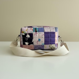 May include: A patchwork crossbody bag with a cream-colored strap. The bag is made of various fabric squares in shades of purple, pink, and blue, with floral and geometric patterns. The bag is sitting on a white surface.