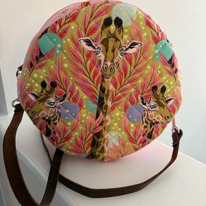 May include: Round, quilted handbag with a giraffe print. The bag features a brown leather strap and a colorful pattern of giraffes, pink leaves, and teal accents on a green background. The bag is approximately 10 inches in diameter.
