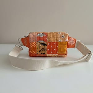 May include: A patchwork fanny pack in shades of orange, featuring various fabric patterns including polka dots, floral designs, and geometric shapes. The bag has a cream-colored adjustable strap and a silver zipper.