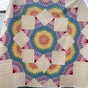 May include: A large quilt featuring a central star pattern composed of small, multi-colored fabric diamonds in shades of pink, yellow, blue, and purple. The star is set against a cream-colored background with visible linear stitching.