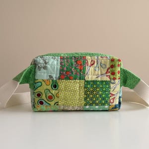 May include: A patchwork fabric bag with a rectangular shape. The bag features a variety of green fabrics with patterns including avocados, scissors, and floral designs. It has a green top and sides, with cream-colored straps extending from each side.