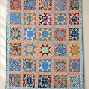 May include: A rectangular patchwork quilt featuring a grid of star-patterned blocks in various colors and fabric prints, including red, blue, yellow, and floral patterns. The quilt is bordered by a light blue fabric with a small floral print.