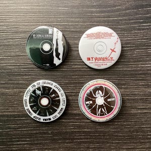 May include: Four black and white CDs with different designs. The first CD has a black and white image of a person hanging from a building with the text "My Chemical Romance" on the top. The second CD has a white background with the text "My Chemical Romance" in red and black. The third CD has a black background with a clock face design and the text "Famous Last Words" and "I Disappear" on the top. The fourth CD has a black and white spider web design with the text "Danger Days" on the top and "MCR" in the center.