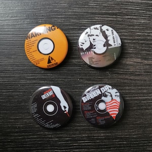 Puede incluir: Cuatro botones redondos de la banda Green Day. Los botones son negros y blancos con un diseño rojo, blanco y azul. Los botones muestran el nombre de la banda y los títulos de los álbumes, incluyendo "Warning", "Dookie", "21st Century Breakdown" y "American Idiot".