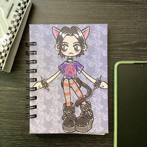 May include: A purple spiral bound notebook with a cartoon illustration of a cat-like person wearing a purple shirt with an anarchy symbol, striped pants, and black boots. The background is a light purple with a repeating pattern of cats.