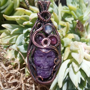 以下が含まれることがあります： 手作りのペンダント。彫刻された紫色のスカルが特徴で、銅線で包まれています。ペンダントには、小さくて丸い紫と透明の宝石が含まれています。背景は、緑色の多肉植物のぼやけたビューです。