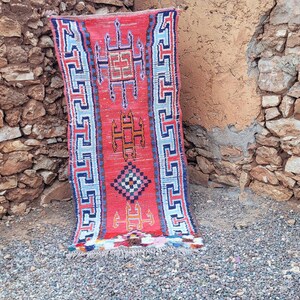 Puede incluir: Una alfombra rectangular roja con un patrón geométrico. La alfombra presenta un diseño central con formas naranjas y negras, enmarcado por bordes azules y blancos. La alfombra está apoyada contra una pared de piedra.