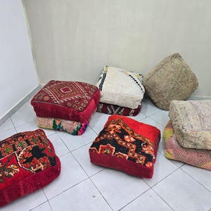 May include: A collection of square floor cushions featuring various woven patterns and textures. The cushions display a range of colors including deep red, black, cream, beige, and muted pink, with some showing intricate geometric and floral designs.