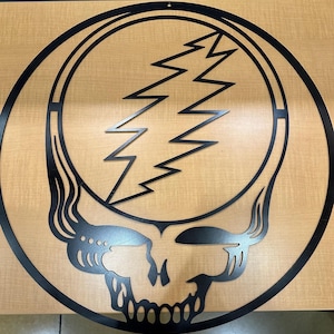 May include: Black metal wall art featuring the Steal Your Face logo, a skull with a lightning bolt. The design is a circular shape with a skull at the bottom and a lightning bolt in the center. The art is mounted on a light brown surface.
